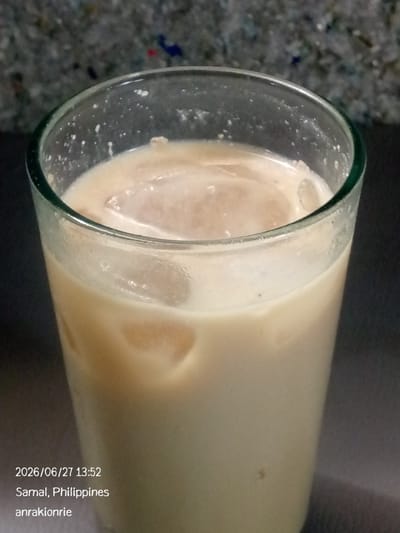 A close-up, eye-level shot shows a glass filled with a light brown liquid and ice cubes. The glass has a green rim. The background is dark and out of focus, with a speckled texture visible at the top. Text overlays the bottom left of the image, indicating a date and time of "2026/06/27 13:52", the location as "Samal, Philippines", and a username "anrakionrie".