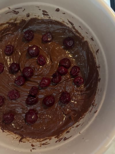 A top-down shot shows a white mixing bowl filled with chocolate batter. Several dark red, wrinkled cherries are scattered on top of the batter. There is no evidence to indicate this image was taken in Kurovskoye, Russia.
