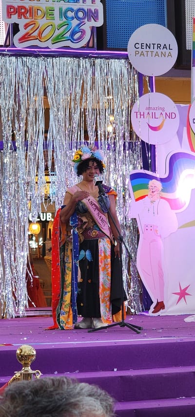 This media file captures a vibrant scene from a Pride event in Baan Chaweng, Thailand. The central subject is a person dressed in an elaborate, colorful outfit, adorned with a floral headdress. They are holding a microphone and appear to be on a stage or platform decorated with shimmering silver fringe.
Behind the individual, a large banner features a stylized drawing of a person with a rainbow accent. To the left of this banner, there are circular signs. One reads "CENTRAL PATTANA," and another says "amazing THAILAND." A prominent sign on the left side of the frame displays "PRIDE ICON 2026" in bold, rainbow-colored letters.
The overall setting is festive and celebratory, typical of a Pride event. The purple stage or backdrop suggests a formal presentation. The assortment of colorful fabrics draped around the central figure adds to the visual richness of the scene. The lighting appears to be artificial, indicating the event is likely taking place indoors or after dark. The atmosphere seems joyful and engaging, with the main subject exuding a confident presence.