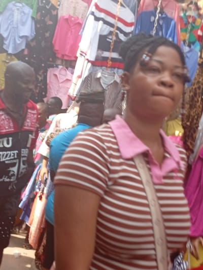 This image depicts an outdoor market scene in Fegge, Nigeria, characterized by numerous clothing items displayed for sale. In the foreground, a young adult female is partially visible, slightly out of focus, facing to her right. She wears a short-sleeved, brown and white horizontally striped polo shirt with a pink collar and has braided dark hair with small, decorative pins near her temples. A light-colored strap crosses her chest diagonally. To her left, in the midground, an adult male with dark skin is seen in profile, smiling. He wears a red polo shirt with black sleeves featuring white text or graphics. Several other individuals are present in the midground and background, largely obscured or blurred. The background is dominated by dense displays of various hanging garments, including shirts and blouses in assorted colors such as pink, white, blue, and patterned black, suspended from what appear to be market stalls or racks. The ground is partially visible, indicating an outdoor, possibly unpaved, surface.