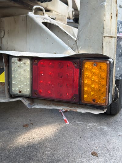 The image shows the rear light assembly of a vehicle, possibly a trailer, which has three distinct sections: a white LED light on the left, a red LED light in the middle, and an amber LED light on the right. The assembly is mounted on a weathered, metallic surface with visible rust and damage. The lights are rectangular with rounded corners and appear to be illuminated by multiple small LEDs. The metallic housing is bent and appears to be detached from the main body of the vehicle on the left side. A piece of red and white striped tape or ribbon is visible on the ground beneath the light assembly, along with some debris. The background consists of a concrete surface, suggesting an outdoor environment. The image is captured from a medium-close, slightly angled perspective, focusing on the rear lights and their immediate surroundings. There are no people visible in the image. The context of Brest, Belarus is not visually inferable from the image itself.