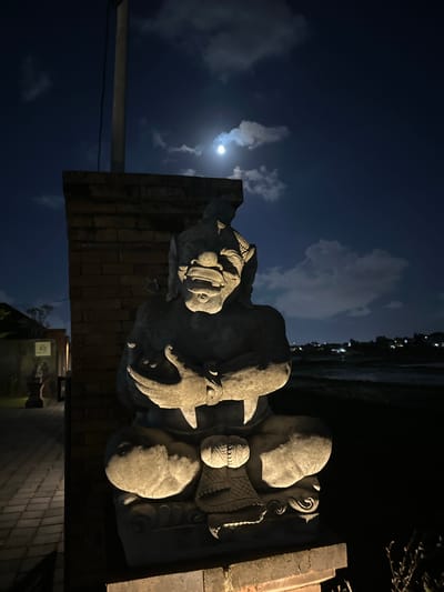 At night in Mengwi, Indonesia, a stone statue of a creature with a distorted face, fangs, and a headdress sits in a meditative pose. The statue is illuminated by artificial light, casting shadows on its surface and highlighting its intricate details. The background is dark, with a sliver of the moon visible through scattered clouds, and a distant line of trees and lights suggesting a populated area. The statue is positioned next to a brick wall and a paved walkway.
