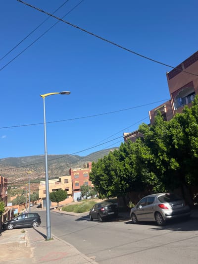 The image depicts a street scene in Ouaouizert, Morocco, under a clear blue sky. A street lamp with a yellow curved arm extends into the frame. Overhead electrical wires crisscross the sky. Buildings, ranging in height and color from peach to yellow, line the street and reach up into the tree-covered hillside in the background. Several cars are parked along the side of the street. The road is paved and bordered by a concrete curb.