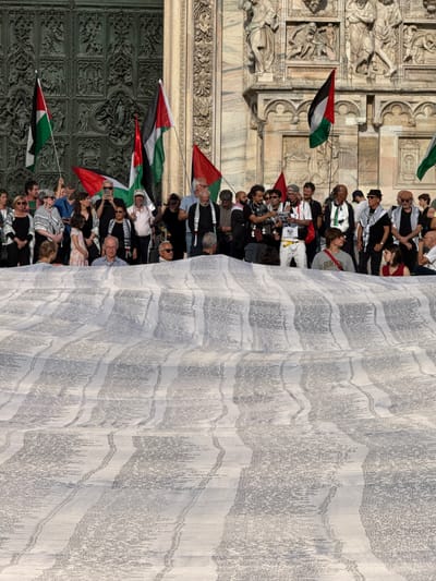 An outdoor gathering is depicted in front of the ornate gothic facade of the Duomo di Milano (Milan Cathedral) in Italy, specifically near one of its large bronze doors. In the midground, a diverse group of approximately 30-40 individuals, primarily adults, is assembled. Many participants are holding Palestinian flags, characterized by horizontal stripes of black, white, and green, with a red triangle at the hoist. Some individuals are wearing keffiyehs draped around their necks. One person in the center-left is speaking into a microphone, while another in the midground-right wears a grey sweatshirt with "BURTON" printed on the chest.
Dominating the foreground and extending across the lower two-thirds of the image is a very large, light-colored fabric banner or sheet, spread horizontally. This banner is densely covered with columns of small, dark text. Closer inspection reveals these to be lists of names, likely in both Arabic and Latin scripts, suggesting a compilation such as a casualty list. The banner is slightly wrinkled, creating subtle variations in its surface. The architectural details of the cathedral, including sculptures, reliefs, and intricate stonework, form the immediate background under bright daylight conditions.