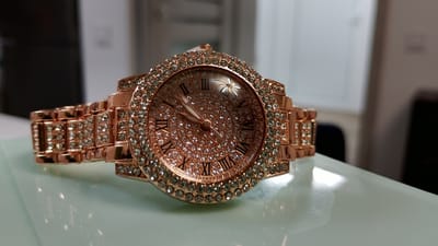 A rose gold, diamond-encrusted watch with Roman numerals rests on a glossy white surface. The watch has a round face with a light-colored dial that features a floral design at the 12 o'clock position. The bezel and band of the watch are covered in small, clear gemstones. The background is out of focus, showing what appears to be an indoor environment with soft lighting. There are no people or animals visible in the image. The location in the city of Smolyan, Bulgaria, is not discernible from the provided image.