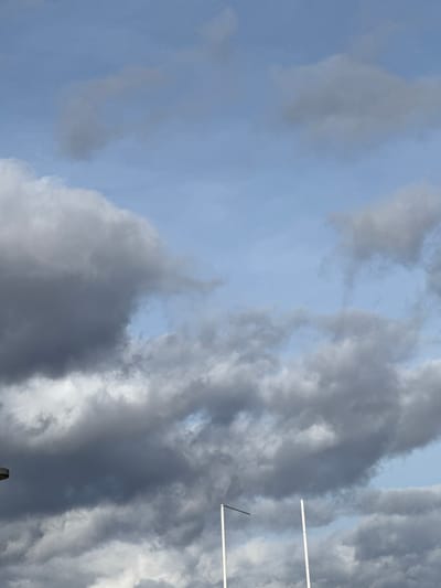 A sky filled with grey and white clouds, obscuring the blue in some areas. On the right side are goalposts, their white bars clearly visible against the textured sky. A dark object is barely discernible at the bottom, contrasting with the light, cloudy backdrop above. The clouds have a rippling texture.
