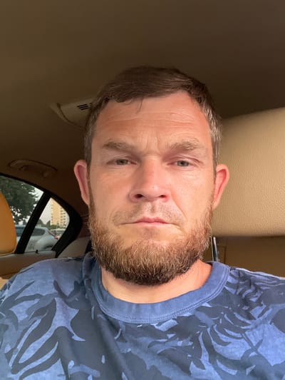 This is a selfie of a man in a car. The man has short, brown hair with graying at the temples and a light beard. He is wearing a dark blue t-shirt with a leafy pattern. His expression appears neutral. The interior of the car is visible, with tan leather seats and the headliner. Through the window, trees and buildings can be seen, suggesting an urban or suburban setting. The lighting suggests it is daytime, possibly afternoon. The image is oriented vertically, making it a portrait-style selfie. No visible text is present in the image. The context provided indicates the location is Krasnodar, Russia.