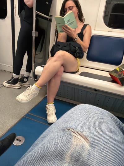 Two individuals are seated on a public transport vehicle, likely a bus or metro, with a blue upholstered seat visible behind one of them. The person in the foreground is partially visible, wearing light blue denim jeans with a noticeable rip in the knee. Their legs are crossed, and they are facing away from the camera.

The second individual, in the mid-ground, is reading a book. They are wearing glasses, a black sleeveless top, and yellow shorts. Their legs are also crossed, and they are looking down at the book. A black bag is slung over their shoulder, and they are wearing white sneakers with yellow-trimmed socks. To their left, another person is standing, wearing black leggings and black high-top sneakers. The interior of the vehicle appears to be utilitarian, with metal poles and white paneling. The floor is a textured blue material.

The scene does not contain any discernible features that would specifically place it in Tbilisi, Georgia.