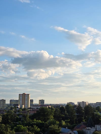 This media file depicts an outdoor scene with a clear blue sky dominating the left half of the frame, dotted with fluffy white clouds. The right half of the image showcases a city skyline with several multi-story buildings of varying architectural styles. Green trees and foliage are interspersed between the buildings, suggesting a city with pockets of nature. Some buildings appear to be residential, with visible windows and balconies. The lighting suggests it is daytime, possibly late afternoon or early evening, as the sky is bright but not harsh, and there's a soft glow to the clouds. The presence of apartment buildings and the general urban landscape indicate a city setting. While the image is taken from an elevated perspective, there are no specific landmarks to pinpoint the exact location within Kyiv, Ukraine, beyond the general urban context. No people or specific activities are visible, and the overall mood is serene and observational. There is no visible text in the image.