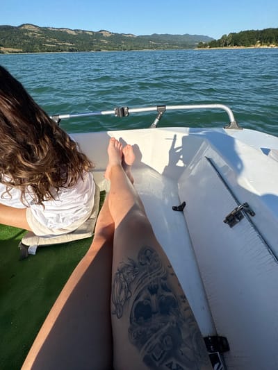 The image captures a point-of-view shot from a boat on a lake, with two individuals on board. In the foreground, the legs of one person are visible, featuring a large tattoo on the thigh. This person is wearing shorts and a white shirt, and is seated on a cushion. The other person's back and long, brown hair are visible, also seated on a cushion. The boat is white with a metal railing. The lake is calm with ripples, and a shoreline with trees and hills is visible in the background under a clear blue sky. The location is identified as the lake in Golyama Zhelyazna, Bulgaria.