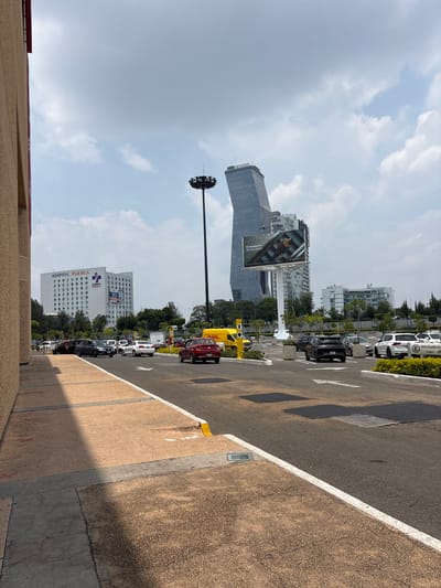 The image captures a daytime urban scene in Puebla, Mexico, under a partly cloudy sky. The perspective is from a slightly elevated position looking down and across a street or parking area.
In the background and to the left, a prominent, modern glass-clad skyscraper with an angular design is visible. Text near its top includes "HP" and part of what appears to be "Inxignia". Below and to its right, a large billboard displays an advertisement featuring a person exercising, with text that reads "El momento de una pausa" (The moment for a pause) and "ANGELOPOLIS". Further right and slightly below the billboard, another multi-story white building is clearly identifiable as "HOSPITAL PUEBLA" with its logo.
In the mid-ground and right side of the image, a paved road or parking lot stretches into the distance, marked with white lines and directional arrows. Several vehicles are present, including a distinctive yellow DHL delivery truck. Another parked car has a visible sign reading "EXCLUSIVO". A tall, dark light pole with multiple fixtures extends into the sky on the left side of the road.
The foreground consists of a light brown concrete barrier or sidewalk, casting shadows onto the darker asphalt surface of the road. The overall impression is an active, modern city environment.