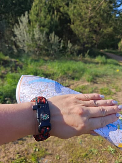 A close-up shot shows a person's left hand holding a map. The map appears to be a tourist or topographical map, showing geographic details such as bodies of water, roads, and possibly terrain features. On the wrist of the arm holding the map, there is a survival bracelet with a compass. The bracelet is made of woven paracord in a red and green pattern and has a black plastic buckle that incorporates a compass. The hand has fingernails that are painted with a light color and a subtle design.
The background is a green, natural environment. There are trees and grassy areas, suggesting a park or wooded area. A path is barely visible in the upper right.
The image doesn't contain enough detail to pinpoint the exact location in the city of Марковский, Russia.