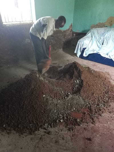 The image shows a man mixing cement in a room with a dirt floor. The room is located at the coordinates 0.545, 34.793 in Lwandeti, Kenya. The man is wearing a white shirt and black pants. He is using a red shovel to mix a pile of brown and gray cement. The cement is mixed with a red liquid. The man is in the center of the image. Behind him is a window with a metal grate. In the background is a couch covered with a white and blue patterned sheet. The floor is dirt, and there is a patch of red liquid on the floor. The walls are painted a pale green and are partially covered in dirt. The room appears to be a simple dwelling. The man is mixing cement to build or repair the structure.