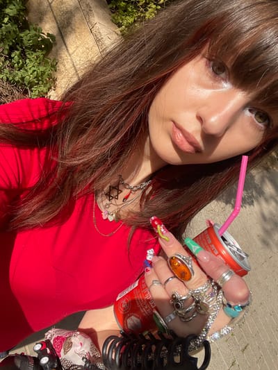 A close-up, high-angle self-portrait features a young individual with long brown hair, reddish highlights, and bangs, looking directly at the camera. She wears a red top and numerous pieces of jewelry, including multiple necklaces with pendants such as a Star of David and a red Hamsa/Evil Eye charm, along with several rings on each finger, including a large amber-colored stone ring on her right ring finger. Her long fingernails are elaborately decorated with various designs, including a green serpentine pattern and dice motifs. She holds two red Coca-Cola cans, visible with Hebrew text; one contains a pink plastic straw. Partially visible near her left side is a floral-patterned fabric, and a black hair claw clip is near her right hand. The background indicates an outdoor setting with green foliage and a light-colored paved surface under bright sunlight, consistent with an urban environment in Raanana, Israel, as evidenced by the Hebrew branding on the cans.