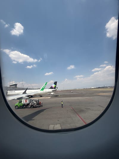 Captured from an airplane window, the image depicts a sunny day at an airport tarmac, with a clear blue sky and scattered white cumulus clouds. The curved outline of the aircraft window frames the scene.
Two passenger jets are visible on the tarmac. The prominent aircraft in the foreground is a white jet with "volaris" written on its fuselage and "VIVA" in green on its tail, which also features a distinctive black and green geometric pattern. Partially visible behind it is another white aircraft, whose tail is black with multi-colored geometric shapes and includes text like "CIA" and "DXA".
A green and white ground support vehicle, marked with "D-189 A" and "ASA 4104", is positioned next to the wing of the Volaris aircraft. The concrete tarmac below is marked with various yellow and red lines. In the distance, a person wearing a yellow high-visibility vest stands on the tarmac, appearing to direct ground operations with outstretched arms. Further in the background, airport buildings and a faint mountainous or urban landscape are visible.
The scene captures typical daytime airport activity, suggesting preparations for departure or arrival. The presence of Volaris and Viva Aerobus aircraft strongly indicates an airport in Mexico, aligning with the provided location context of Mexico City, Mexico.
