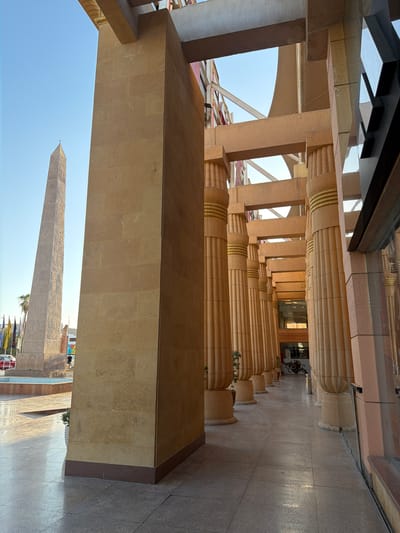 The image depicts a view of the exterior of a building with a long colonnade of tall, fluted pillars made of beige stone. The pillars are adorned with gold bands at intervals. Above the pillars, there are architectural elements with hieroglyphic carvings. To the left of the pillars, the clear blue sky is visible, with hints of a white canopy or roof structure and diagonal supports.
In the foreground, a large, rectangular beige stone structure extends horizontally across the lower portion of the image. Below this structure, a bright blue sky is visible, and in the distance, an ancient Egyptian obelisk with hieroglyphs is prominently featured. The obelisk stands on a paved area, with flags and some vehicles visible at its base, suggesting a public or tourist area. The overall setting appears to be an outdoor architectural space, likely a historical or themed attraction, given the presence of the obelisk and hieroglyphs.
The lighting suggests it is daytime, with bright sunlight casting shadows. The weather appears clear and sunny. The scene evokes a sense of ancient grandeur and historical significance. No people are clearly discernible as distinct subjects, though the presence of vehicles and flags implies human activity in the vicinity. The location context of Sharm El Sheikh, Egypt, further supports the interpretation of ancient Egyptian architectural elements and a tourist destination. There is no discernable text in the image, apart from the hieroglyphs on the obelisk and architectural elements.