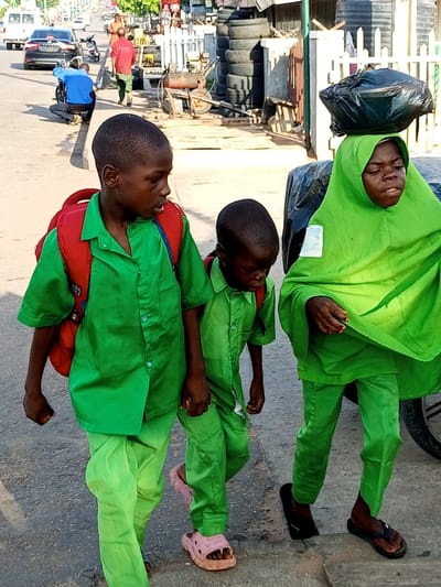In Tempure, Nigeria, three children dressed in bright green uniforms walk on a paved street. Two boys, one in the foreground and one behind him, wear red backpacks over their green shirts and trousers. The boy in the foreground has a serious expression. To their right, a girl wearing a green hijab, also in a green uniform, carries a large black plastic bag balanced on her head. The background shows a busy street scene with vehicles, buildings, and other people. The overall atmosphere is one of daily life and activity on a Nigerian street.