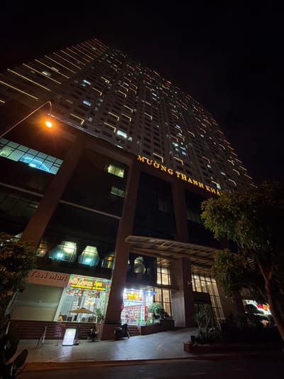 The image captures a nighttime view of a tall, modern building illuminated by numerous yellow lights along its exterior. The building is adorned with vertical rows of lights and also has lights on many of its windows, suggesting it is occupied or has active businesses. One side of the building displays large, yellow lettering that reads "MUONG THANH KHA".
Below the towering structure, the ground level features shops and commercial establishments. One prominent sign, illuminated in bright colors, says "Cogna Spa" with additional text in Vietnamese. Another sign visible on the building's facade reads "Tan Mart". The entrance to these establishments is brightly lit, showcasing a variety of products and displays, likely indicating retail activity.
The overall scene is set at night, with the darkness of the sky contrasting with the artificial lights from the building and the businesses below. Trees with lush green foliage are visible, adding a natural element to the urban setting. The lighting from a streetlamp is also visible in the foreground, casting a warm glow. The image seems to depict a lively commercial area in Nha Trang, Vietnam, at night.