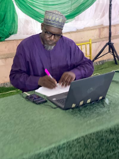 A man is sitting in front of a laptop, writing on a piece of paper with a pink marker. He is wearing a purple tunic and a patterned cap. The man has glasses and a grey beard. He appears to be focused on his task.

The scene is outdoors, as suggested by the green, artificial turf that covers the ground. Behind him, there are draped green and white fabrics, possibly part of a decoration or backdrop for an event. A yellow railing can be seen in the background, and a black tripod stands behind the man's head. A laptop is open on a surface in front of him, and a mobile phone is visible next to it.

The lighting suggests it might be daytime, but the exact time is unclear. There is no visible text in the image. The overall atmosphere seems to be one of quiet concentration during an event or meeting. The setting in Unguwar Sarki, Nigeria, provides a cultural context for the attire and possibly the event.