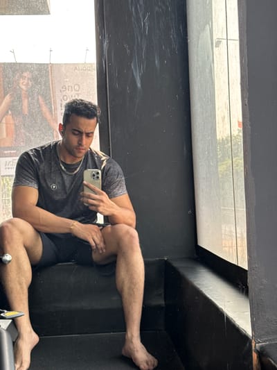 A man is taking a selfie in a mirror. He is wearing a t-shirt, shorts, and a necklace. He is sitting with his legs crossed. The reflection in the mirror reveals an exterior with urban elements that are consistent with those found in Kolkata, India. The reflection includes what appears to be a billboard with text and images on it, as well as indistinct buildings in the background. The mirror is situated in a structure of some kind.