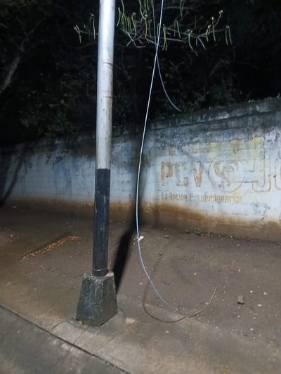 This nighttime image depicts an outdoor scene in Altagracia, Venezuela, showing an urban or semi-urban environment. A prominent metal lamp post stands slightly left of the image's center in the foreground. Its upper section is painted a light grey, and the lower section is dark grey or black, with visible rust or wear at the transition. The post is anchored to a weathered, roughly octagonal concrete base exhibiting cracks and discoloration. A thin, light-colored cable or wire hangs from the upper portion of the lamp post, curving downwards to rest on the unpaved ground below.
Behind the lamp post, a concrete block wall extends horizontally across the middle ground. The wall's original light-colored paint is heavily faded and discolored, with a noticeable brown stain along its base and patches of grey and blue. Distinct, faded orange-brown graffiti is present on the wall, spelling "PCV" alongside a stylized rooster emblem. Below this, the slogan "La Opción Revolucionaria" (The Revolutionary Option) is also visible in faded orange-brown text.
The ground in the foreground consists of a concrete sidewalk on the left, transitioning into an unpaved dirt area around the lamp post and extending to the wall. This dirt area contains small stones and debris. Dark trees and foliage are visible in the background, above and behind the wall, with several long, thin, light-green tendrils hanging from branches directly above the lamp post. The scene is dimly lit, casting shadows from the lamp post onto the wall and ground, suggesting an external light source from the right. No individuals or specific actions are depicted within the scene.