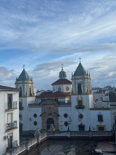 An elevated, daytime view captures the Church of Nuestra Señora de la Merced in Ronda, Spain, beneath a partly cloudy sky. The prominent central structure features a white stucco facade with yellow ochre trim. It includes two symmetrical bell towers topped with pyramidal roofs adorned in a blue, white, and ochre chevron tile pattern, each surmounted by a decorative finial or cross. A central, reddish-brown tiled dome is topped by a smaller white cupola and a cross. The church's main entrance displays an ornate, classical-style portal with a statue of a robed figure positioned within a niche above the doorway.

In the immediate foreground, a dark, flat rooftop, possibly covered with a tarp, appears wet and reflects ambient light. A low, gray concrete parapet wall defines the edge of this rooftop. Several large, ornate chandelier-style decorative light fixtures, composed of gold and white elements, are suspended at various heights between the foreground rooftop and the church facade. To the left, a multi-story, white residential building with dark balconies and an external air conditioning unit partially obscures the left bell tower. The background reveals a dense urban landscape of white-washed buildings with reddish-brown tiled roofs, typical of a Spanish town, receding into the distance under the soft light. No individuals are visible, and no overt actions are occurring.