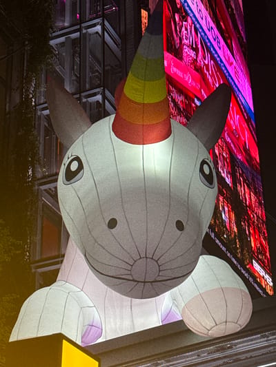 An illuminated, inflatable unicorn head is displayed prominently in front of a blurred, vibrant digital billboard in an urban setting. The unicorn, rendered in shades of white and gray, features a rainbow-colored horn, large cartoonish eyes with black pupils and gray irises, and small, dark oval nostrils. Its body appears to be that of a plush toy, with rounded ears and a soft, rounded snout. The unicorn's head is lit from within, casting a soft glow.
Behind the inflatable, a massive LED screen displays colorful, animated advertisements. The billboard shows vibrant pink and red hues, with text in English that is partially obscured by the unicorn. One visible phrase reads "destination for world-class entertain". The background also shows architectural elements of a building, possibly a skyscraper, with dark glass and metal framing, and some dark foliage or vines on the left side. The scene is dimly lit, suggesting it is nighttime.
The digital billboard and its surrounding structures are characteristic of the illuminated advertising displays found in areas like the Ratchaprasong intersection in Bangkok, Thailand, a well-known commercial and entertainment hub with numerous large shopping malls and prominent digital billboards. The presence of such a large, whimsical inflatable in a high-traffic urban area is consistent with promotional events or installations often seen in prominent Bangkok districts.