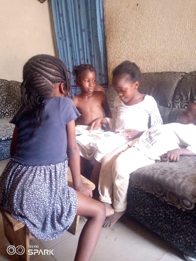 An indoor scene in Ajaokuta, Nigeria, depicts four children within a room featuring a textured light-colored wall and blue patterned curtains. A dark-patterned sofa with multiple cushions is positioned on the right side of the frame. In the foreground, a child with braided hair, wearing a dark blue t-shirt and a blue and white floral patterned skirt, sits on a small wooden stool, facing away from the camera. Adjacent to her, a second child, bare-chested and wearing light-colored lower garments, sits and observes the activity to her right. A third child, wearing a white patterned long-sleeved top and light-colored trousers, sits on the sofa, intently looking down at several white rectangular objects (possibly cards or a book) held in her lap, with her right hand positioned over them. To the right of the third child, a fourth child is partially visible, lying reclined on the sofa, dressed in a light-colored top with dark graphics and light-colored trousers, a pink band visible on her left wrist. The two central children appear to be interacting with the objects in the third child's lap. The floor is a light-colored solid surface.