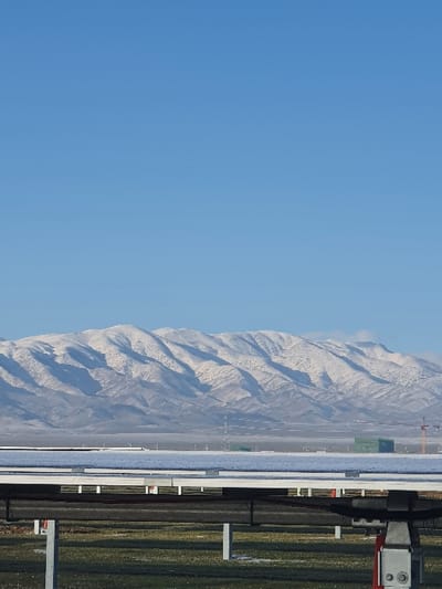 A landscape view features a clear, light blue sky occupying the left portion of the frame. Moving right, a mountain range, dusted with snow, stretches towards the background. In the foreground, a fence or barrier of some sort runs vertically, with linear elements and supports visible against a backdrop of green vegetation. The scene presents a blend of natural and man-made structures.