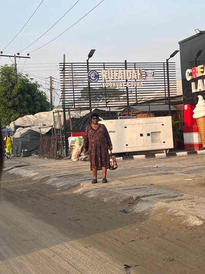 An adult female, dark complexion, stands centrally on a dirt and concrete roadside in Lekki, Nigeria, looking towards the viewer. She wears a long-sleeved, V-neck brown dress with a subtle pattern, dark sunglasses, and dark flat shoes. She holds a floral-patterned tote bag in her right hand. To her left, another individual in a bright yellow top walks away from the camera. The road exhibits tire tracks and is bordered by a concrete kerb and gutter. A small bird is visible on the road surface near the bottom.

Behind the woman, a large white industrial generator, labeled "MIKANO," rests on a raised platform. To the generator's right, a dark commercial structure features partially visible "CREAM LAND" lettering and a large, decorative ice cream cone sculpture. Further left, a series of makeshift market stalls or structures, constructed from dark tarpaulins and metal frames, are visible, alongside piles of bundled materials.

In the background, a large billboard with horizontal black bars displays "RUFAIDAH YOGHURT & FURA" in white text, accompanied by a yogurt cup logo. Overhead, power lines crisscross a clear, pale blue sky. Green foliage from trees is visible behind the market stalls and below the billboard.