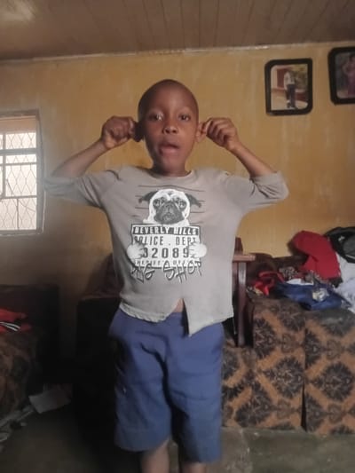 In Yelwa, Nigeria, a young boy stands in a room, his hands cupping his ears. He wears a light gray t-shirt with a graphic of a pug and text that reads "BEVERLY HILLS POLICE DEPT 32089 SHERIFF OF BIG SHOT." He is also wearing blue shorts. The room has yellow walls, a wooden ceiling, and a window with a diamond pattern on the left. Two framed pictures hang on the wall to the right. In the background, there are two patterned couches, one with clothes draped over it.
