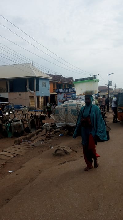 This image depicts a bustling street scene in Ajaokuta, Nigeria, under an overcast sky. In the foreground, a woman walks purposefully towards the viewer, positioned slightly to the right of the frame's center. She wears a teal head covering and a loose-fitting, long teal garment over red trousers, with a small bag slung across her chest. Demonstrating a common local practice, she expertly balances a large, light-colored rectangular container on her head.
The street is a mixture of unpaved, dusty ground on the left, transitioning to a more compact, darker surface in the immediate foreground and where the woman walks. The left side of the street is cluttered with various objects, including several wheelbarrows, stacks of blue pipes, wooden planks, and other materials, some covered with light-colored tarpaulins, suggesting a market or construction supply area.
In the mid-ground and background, numerous other people are visible, walking, standing, and interacting. Many appear to be men dressed in traditional attire. Several multi-story buildings line the street; prominently featured are a light-blue building with a white roof on the left and an orange-brown building further back. Overhead power lines stretch across the cloudy sky.
A clear sign on a shop or stall in the mid-ground displays the text "RUFAIDAH YOGHURT," indicating a business. A small, partially visible yellow auto-rickshaw is seen on the far right, alongside a street light pole. The scene conveys the lively atmosphere of a typical day in a Nigerian town.