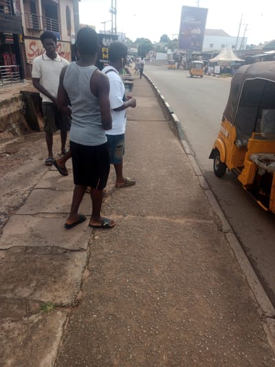 The image shows a street scene in Enugu, Nigeria. Three young men are standing on a sidewalk. The man on the left is wearing a white polo shirt and dark shorts. The man in the middle is wearing a grey tank top and black shorts. The man on the right is wearing a white t-shirt and denim shorts. They are standing next to a road with a yellow tricycle, commonly known as a "keke," parked on the side. The background shows buildings and a billboard advertising a Cadbury product. The sidewalk is made of concrete and asphalt. The overall lighting suggests it is daytime.