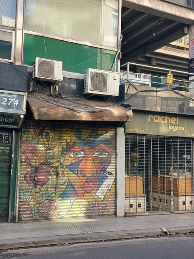 The image depicts the storefront of "rachel joyeros" located at Libertad 278 in Buenos Aires, Argentina. A metal security gate with an abstract, colorful mural covers the lower portion of the storefront. The upper portion of the shop is protected by a metal grid. Above the mural, the shop's name and address are visible on a dark green sign in gold lettering. Air conditioning units are mounted on the exterior wall to the left of the storefront, interspersed with a tangle of wires. A portion of the adjacent building at number 274 is visible on the lower left. The street and sidewalk are visible to the right.