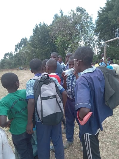 The image shows a group of 7 young boys in a forest clearing near Malava, Kenya, located at coordinates latitude: 0.53, longitude: 34.81.  The boys are dressed in school uniforms, and one is wearing a backpack. The group is standing in a circle, and one of the boys is holding a white object, possibly a plastic bottle. The forest behind them is dense and green. The ground is covered in grass. The boys are engaged in a conversation. There are traces of dirt and brown grass scattered throughout the clearing.