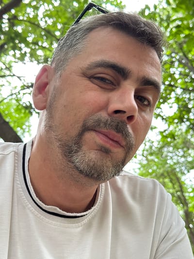 A close-up, low-angle, outdoor selfie captures the upper body and face of a man with short, graying brown hair and a goatee. He wears a white t-shirt with black and white striped trim around the neckline. Sunglasses are perched on his head. The background is a bokeh of green foliage, suggesting he is outdoors, likely in a park or natural setting. The lighting is bright, indicating daylight. No specific location in Almaty, Kazakhstan can be determined from the image.