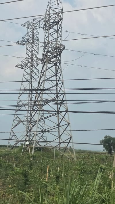 The image shows a power transmission tower situated in a rural area, likely near Xuân Long, Vietnam. The tower is made of steel and has a lattice structure with numerous cross-braces. Electrical wires are suspended from the tower. The surrounding area appears to be a field with vegetation. The sky is overcast.
