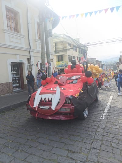 The image depicts a street scene in Amaguaña, Ecuador, during what appears to be a parade or festive event. The street is paved with dark cobblestones, from which grass is growing in the interstices.
The central subject is a dark red sedan, likely a Volkswagen, extensively modified and decorated to resemble a monster or dragon. The front bumper features large, prominent white pointed "teeth," and the hood and parts of the sides are covered with red fabric adorned with black eye-like decals. A black cape-like fabric is draped over the driver's side front fender and wheel. The car's roof supports multiple clusters of black, red, and gold balloons, along with a red and black circular arched structure from which a small white garment hangs.
On the left side of the street, a two-story, light-colored building with traditional wooden-framed windows is visible. A street number sign "Oe1-18" is attached to its facade. The ground floor houses a retail establishment displaying numerous white athletic shoes in a glass cabinet. A utility pole, laden with a dense network of overhead electrical and communication wires, stands between the building and the street, also featuring a "WiFi libre" sign and a partial street sign reading "A VIA".
Several individuals are present. In the foreground, on the left sidewalk, a woman with dark hair, wearing a black t-shirt and dark pants, walks past the shoe store while holding a dark object, likely a mobile phone. Another woman in a light purple jacket and blue jeans stands near the decorated car on the left. To the right and behind the decorated car, a procession of people, including multiple children, is observed. The children are dressed in various festive or traditional attire, some in white dresses with colorful ribbons and distinct headwear, and at least one child in a red costume. An adult woman in a blue jacket and light pants walks alongside the children on the right. Other adults are visible further back within the procession and near the surrounding buildings.
Colorful triangular pennants are strung across the street overhead. The background reveals additional multi-story buildings lining the street, with distant hills or mountains visible under a bright, slightly hazy sky.