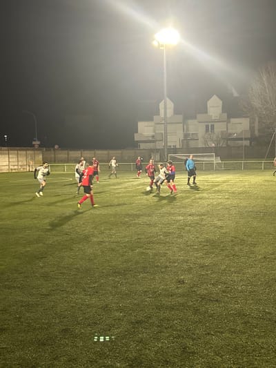 A nighttime soccer match unfolds under bright stadium lights. Two teams, one in red and the other in grey, are in close proximity on a green field, seemingly mid-play. Buildings stand in the background, partially obscured by the night and the light from the stadium. The illuminated soccer field casts long shadows of the players.