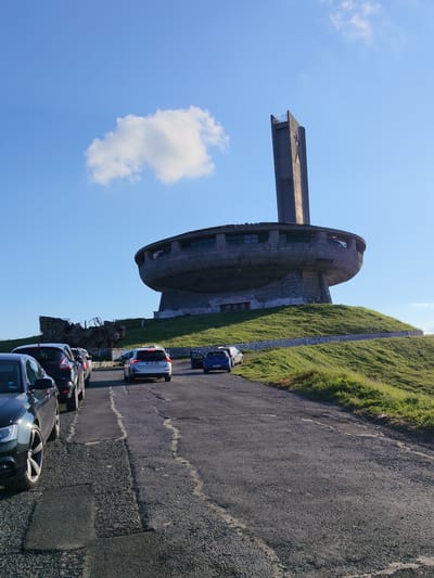The image captures a view of the Buzludzha Monument, a former Communist Party headquarters, situated on Buzludzha Peak in Shipka, Bulgaria. The monument, a massive concrete structure resembling a flying saucer with a tall tower, stands atop a grassy hill under a clear blue sky with a single white cloud. Several cars are parked on a paved road leading towards the monument, with some appearing to be in a queue. The road surface is cracked and weathered. The monument itself shows signs of disrepair, with visible damage to its outer shell and the tower. The surrounding landscape is characterized by green, undulating hills. The overall scene suggests a historical site that is now a tourist attraction, with visitors arriving by car.