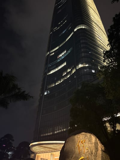 A low-angle shot captures a modern skyscraper at night in Haizhu District, China. The building's facade is characterized by its curved design and vertical lines of illumination, creating a striking visual effect against the dark sky. The warm yellow light emanating from the skyscraper's windows and architectural features suggests an active urban environment.
In the foreground, a large, textured rock with prominent golden Chinese characters carved into it is partially visible. The characters appear to be ornamental or signage, adding a cultural element to the scene. Dark silhouettes of trees frame the upper and lower parts of the image, their branches contrasting with the smooth lines of the building. The overall mood is one of contemporary elegance and nighttime city life. The scene is set at night, with no specific weather conditions noted, and the location is identified as Haizhu District, China. No people are visible.