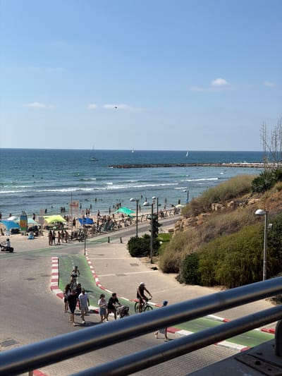 The image depicts a sunny day at a beach in Tel-Aviv, Israel. The scene is divided into the foreground and background. In the foreground, a paved pathway with a red and white striped edge runs diagonally across the image. Several people are walking and cycling on this pathway. A group of four people are walking towards the viewer, with one person pushing a stroller. A woman on a bicycle is heading away from the viewer. Further in the foreground, a railing suggests a viewing platform or balcony.
The middle ground shows a sandy beach populated with numerous people enjoying the sun and sea. Beach umbrellas and tents are set up, and bicycles are parked along the pathway bordering the beach. Surfers are visible in the water, and the sea is calm with gentle waves.
The background consists of the blue sea meeting a clear blue sky. Sailboats can be seen on the horizon, along with a breakwater made of rocks. A small, dark object, possibly a kite or a bird, is visible in the sky. The overall impression is a vibrant and active beach scene on a pleasant day.