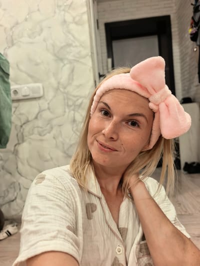 A young woman with blonde hair, wearing a pink plush headband with a large bow and a white pajama top with a subtle heart pattern, is shown in a close-up selfie. She is indoors, likely in a bathroom or bedroom, with a marble-patterned wall and a closed door visible in the background. The lighting suggests it could be evening or a dimly lit indoor space. The woman has a relaxed and pleasant expression, looking towards the camera with a slight smile. Her hand is lightly touching her neck, adding to the casual and comfortable demeanor. The overall mood is one of self-care and relaxation. There is no visible text in the image.