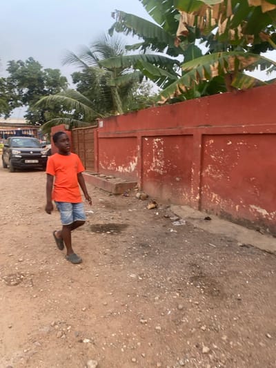 A young person in an orange shirt and denim shorts stands on a dirt road, wearing flip-flops. Behind them is a red-painted wall and a concrete curb, alongside scattered debris. A black car is visible in the background near what appears to be a market area. Tropical trees with large green leaves are to the left, reaching toward the sky. The sky is overcast.