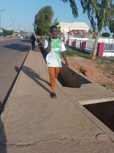 In the city of Tumfure, Nigeria, a young Black male, dressed in a green and white striped t-shirt with the word "HOFMANN" visible and orange pants, walks along a concrete sidewalk next to a road. He is holding a mobile phone to his left ear and appears to be engaged in a conversation. His shadow stretches out behind him on the sidewalk, indicating a sunny day. The road to his left shows moderate traffic, with several vehicles and a motorcyclist visible. Beyond the road, the urban environment includes buildings, streetlights, and trees. To the right of the sidewalk, there is a ditch and a red and white fence with flags. The overall scene depicts a typical street view in an African city on a clear day.