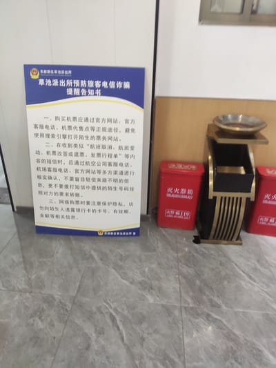This is a medium-shot, eye-level, indoor photograph taken in Jianyang City, China. The image features a blue and white poster with Chinese text, which appears to be a public announcement or notice. To the right of the poster, there is a decorative trash receptacle with gold and black accents and an ashtray on top. Two red fire extinguisher boxes are placed on either side of the receptacle, also featuring Chinese text and the number "119", which is China's emergency telephone number for fire services. The flooring is a gray marble-patterned tile. The overall scene suggests a public indoor space such as a transportation hub, an office building lobby, or a public service area.