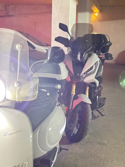 This image depicts a static indoor or covered parking environment in Tangier, Morocco. The scene is illuminated by artificial light sources, casting a warm, orange-toned glow on the background walls and a concrete pillar.
In the foreground, two motorcycles are parked side-by-side. To the left, partially obscured, is a light grey scooter featuring a clear, slightly yellowish windscreen mounted with two visible bolts. The scooter has a black, diamond-quilted seat and "Cappucino" (or similar cursive text) is visible on its left side fairing. The scooter's front includes a visible turn signal with a bright internal light source.
To the right, positioned slightly behind the scooter, is a white and black Honda X-ADV adventure scooter/motorcycle. It is equipped with prominent LED headlights, a dark windscreen, and black handguards on the handlebars. The front suspension consists of gold-colored inverted forks, above a spoked front wheel with a visible disc brake system. The vehicle exhibits a robust stance typical of adventure models.
In the background, behind the motorcycles, a white car is partially visible, parked against a light-colored wall. No individuals are present in the frame. The floor is a dark, rough-textured surface. A vertical concrete pillar stands between the white car and the Honda motorcycle, showing signs of wear or uneven paint.