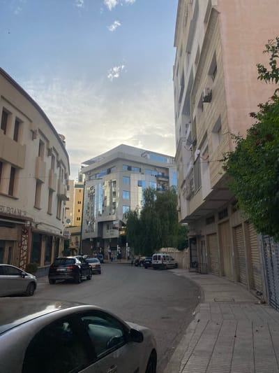 A street scene in Meknes, Morocco, featuring buildings with distinct architectural styles. The sky is partly cloudy with visible sunlight. Buildings of varying heights line the street, including a modern glass-fronted structure. Cars are parked on the side of the street. A tree provides foliage and partial shade in the distance. Architectural details on the buildings include textured facades and protruding elements. Sidewalks and pedestrian areas are present.
