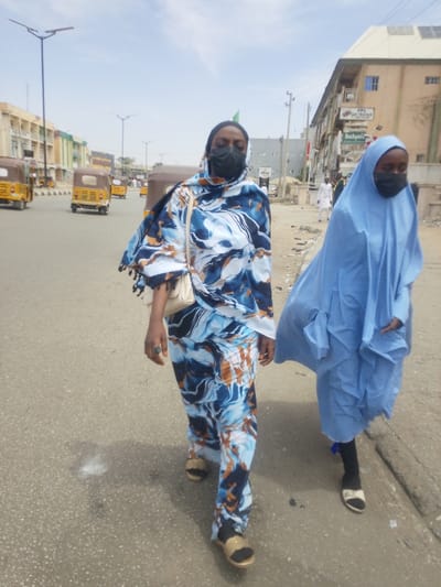 The image depicts a sunlit urban street in Babban Giji, Nigeria. Two females are walking towards the viewer on the paved asphalt road. The female on the left, an adult, wears a black face mask, a patterned headscarf and a matching full-length garment in blue, white, and brown abstract designs. She has gold-colored open-toed sandals and carries a light-colored shoulder bag. A ring is visible on her left ring finger. The female on the right, appearing younger, wears a black face mask, a light blue full-body covering (abaya/niqab style), and black socks with white/cream open-toed sandals. She walks slightly behind and to the right of the lead female.

In the background, the road features multiple yellow and brown three-wheeled auto-rickshaws (Keke Napep/Bajaj RE) on the left side, indicating traffic. Tall street lights with solar panels are positioned along the road and in a central median. Multi-story commercial buildings line both sides of the street. Those on the left have light-colored facades. The building on the right has a light brown facade with solar panels on its roof and displays commercial signs, including 'Buchymix' and 'PPL SHOP RENTAL'. Utility poles are also visible on the right. A male figure in a white robe is seen walking further down the street on the right. Shadows are cast on the ground, consistent with a clear, sunny day.