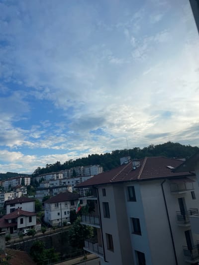 The image presents a view of a town nestled in a valley, with buildings cascading down a hillside under a partly cloudy sky. The architecture suggests a European setting, with terracotta-tiled roofs and a mix of apartment buildings and older, smaller homes. A Bulgarian flag is visible on a balcony of one of the buildings, suggesting the location is in Bulgaria. The foreground features a modern apartment building with balconies. The background is dominated by a lush green hillside covered in trees, indicating a natural, possibly mountainous, environment. The lighting suggests it is either early morning or late afternoon, with soft sunlight filtering through the clouds. The overall impression is one of a peaceful, residential area. The provided metadata states that this image was taken in Zlatograd, Bulgaria.